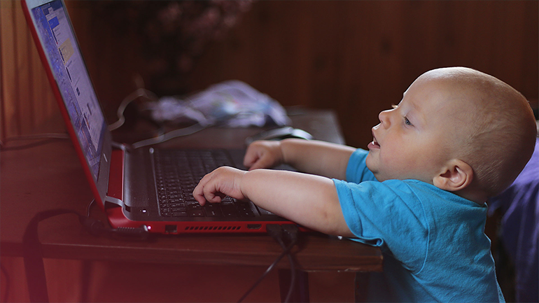 A képen egy kisbaba látható aki alig ér fel a számítógép billentyűzetéig.